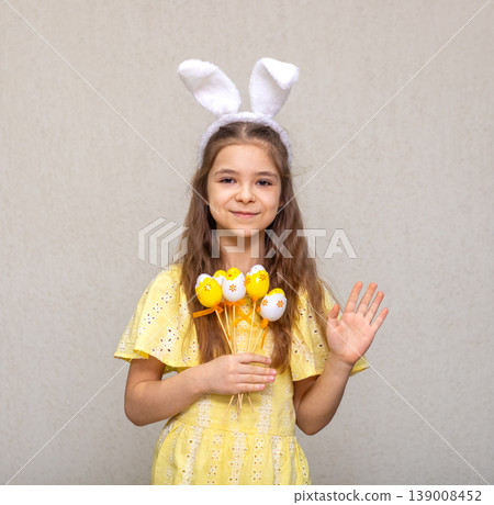 Young girl wearing bunny ears and yellow dress holding decorated Easter eggs and waving 139008452
