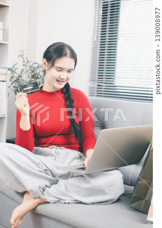 Young Asian woman using laptop and credit card for online shopping at home, browsing ecommerce deals on sofa with shopping bag nearby, digital payment, cashless purchase and retail lifestyle concept Young Asian woman using laptop and credit card for online shopping at home, browsing ecommerce deals on sofa with shopping bag nearby, digital payment, cashless purchase and retail lifestyle concept 139008977
