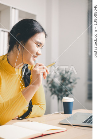 Young Asian woman studying online at home office, wearing earphones and speaking to laptop while holding pen. Remote learning, virtual class, webinar, tutoring and digital education concept. 139009046