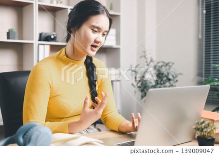 Young Asian woman speaking during online class on laptop at home office desk, learning remotely with expressive hand gestures. Webinar, video call, e-learning, communication and study concept. Young Asian woman speaking during online class on laptop at home office desk, learning remotely with expressive hand gestures. Webinar, video call, e-learning, communication and study concept. 139009047