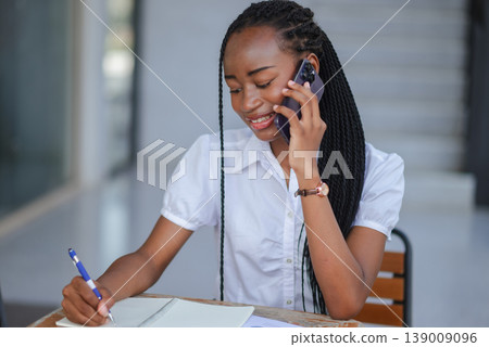 Young African professional woman smiling during a phone call while writing in notebook, concept of customer communication, remote work, planning and modern mobile business productivity. 139009096