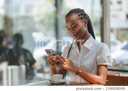 Young African woman sitting at cafe table and using smartphone beside a cup of coffee, concept of digital lifestyle, online communication, remote work and modern business routine. Young African woman sitting at cafe table and using smartphone beside a cup of coffee, concept of digital lifestyle, online communication, remote work and modern business routine. 139009099