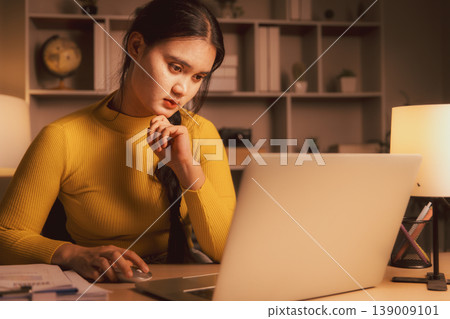 Young Asian woman using laptop and mouse in cozy home office at night, showing focused remote work, online research, problem solving, concentration, analysis and evening productivity. Young Asian woman using laptop and mouse in cozy home office at night, showing focused remote work, online research, problem solving, concentration, analysis and evening productivity. 139009101