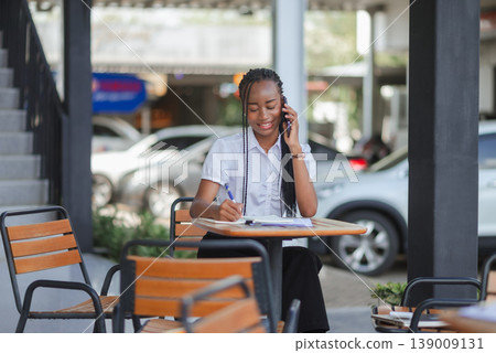Young African business woman talking on smartphone while writing notes at an outdoor cafe table, concept of client communication, remote work, multitasking and modern business lifestyle. 139009131