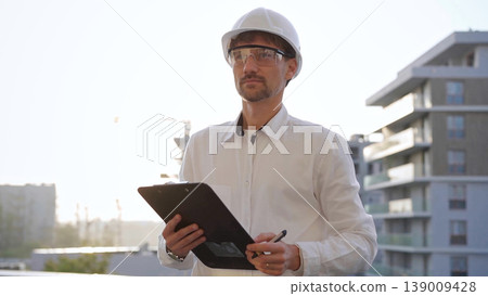 Man construction engineer wearing white shirt and hard hat is making notes on a clipboard while inspecting a building site at sunset, front view. Architecture and engineering concepts Man construction engineer wearing white shirt and hard hat is making notes on a clipboard while inspecting a building site at sunset, front view. Architecture and engineering concepts 139009428