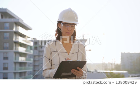 Woman construction engineer wearing white checked blouse and hard hat is making notes on a clipboard while inspecting a building site at sunset, front view. Architecture and engineering concepts Woman construction engineer wearing white checked blouse and hard hat is making notes on a clipboard while inspecting a building site at sunset, front view. Architecture and engineering concepts 139009429