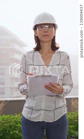 Woman a construction engineer wearing white checked blouse and hard hat is making notes on a clipboard while inspecting a building site in foggy weather, front vertical view. Architecture concept Woman a construction engineer wearing white checked blouse and hard hat is making notes on a clipboard while inspecting a building site in foggy weather, front vertical view. Architecture concept 139009437