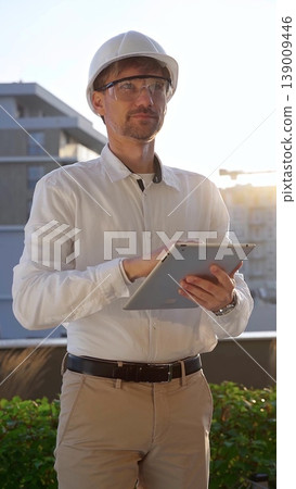 Male construction engineer wearing white shirt and helmet is using digital tablet while inspecting a building site at sunrise, front and vertical view . Architecture concept 139009446