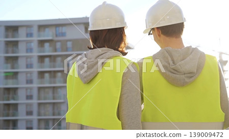 Man and woman architects or engineers wearing safety hard hats and vests holding blueprint and discussing something on construction site at sunrise, back view. Architecture and engineering Man and woman architects or engineers wearing safety hard hats and vests holding blueprint and discussing something on construction site at sunrise, back view. Architecture and engineering 139009472