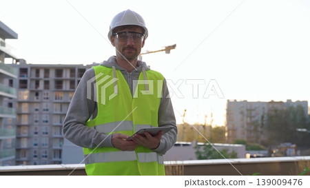 Man construction site engineer holding tablet and wearing hard hat and safety vest with a sunrise at the background in early morning Man construction site engineer holding tablet and wearing hard hat and safety vest with a sunrise at the background in early morning 139009476