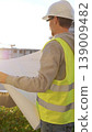 Man an architect or engineer wearing safety hard hat and vest holding blueprint and looking to construction site at sunrise, back vertical view. Architecture and engineering 139009482