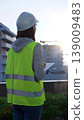 Woman construction engineer with a white hard hat and safety vest is using digital tablet while inspecting a construction site in the early morning at sunrise, back view 139009483