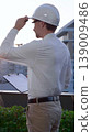 Man a constructive engineer wearing white shirt and hard hat is making notes on a clipboard while inspecting a building site at sunrise, back and vertical view . Architect at work 139009486
