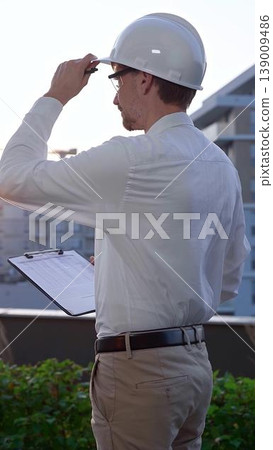 Man a constructive engineer wearing white shirt and hard hat is making notes on a clipboard while inspecting a building site at sunrise, back and vertical view . Architect at work Man a constructive engineer wearing white shirt and hard hat is making notes on a clipboard while inspecting a building site at sunrise, back and vertical view . Architect at work 139009486