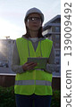 Female architect or engineer wearing safety hard hat and vest holding digital tablet while inspecting a construction site at sunrise in early morning, front vertical view. Architecture concept 139009492