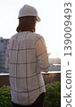 Woman a construction engineer wearing white checked blouse and hard hat is using tablet computer while inspecting a building site at sunset, back and vertical view. Architecture concept 139009493