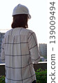 Female construction engineer wearing white checked blouse and hard hat is using tablet computer while inspecting a building site at sunset, back and vertical view. Architecture concept 139009494