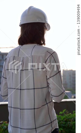 Female construction engineer wearing white checked blouse and hard hat is using tablet computer while inspecting a building site at sunset, back and vertical view. Architecture concept 139009494