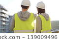 Man and woman engineers wearing safety hard hats and vests holding blueprint and discussing something on construction site at sunrise, back view. Architecture and engineering concept 139009496