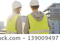 Two architects wearing safety hard hats and vests holding blueprint and discussing construction site project at sunrise, back view. Architecture, engineering and teamwork concept 139009497