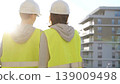 Man and woman architects or engineers wearing safety hard hats and vests holding blueprint and discussing something on construction site at sunrise, back view. Architecture and engineering 139009498