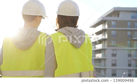 Man and woman architects or engineers wearing safety hard hats and vests holding blueprint and discussing something on construction site at sunrise, back view. Architecture and engineering Man and woman architects or engineers wearing safety hard hats and vests holding blueprint and discussing something on construction site at sunrise, back view. Architecture and engineering 139009498