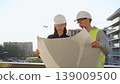 Two architects wearing safety hard hats are holding blueprint and discussing something on construction site at sunrise. Architect and teamwork concept 139009500