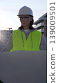 Female architect or engineer wearing safety hard hat and vest holding blueprint while inspecting a construction site at sunrise in early morning, front vertical view. Architecture concept 139009501