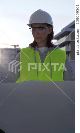 Female architect or engineer wearing safety hard hat and vest holding blueprint while inspecting a construction site at sunrise in early morning, front vertical view. Architecture concept 139009501