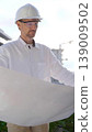 Construction site manager, wearing helmets and safety glasses, is holding blueprints and inspecting a building site during sunrise or sunset, front view vertical portrait. Architecture and engineering 139009502