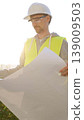 Man an architect or engineer wearing safety hard hat and vest holding blueprint and looking to construction site at sunrise, front vertical view. Architecture concept 139009503