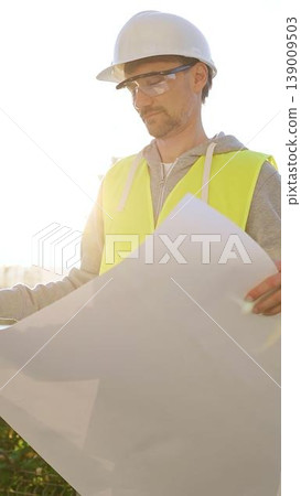 Man an architect or engineer wearing safety hard hat and vest holding blueprint and looking to construction site at sunrise, front vertical view. Architecture concept 139009503