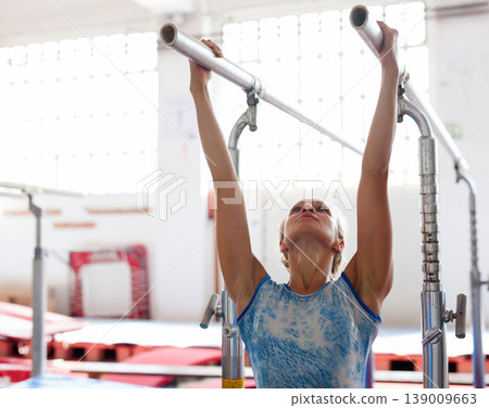 Woman gymnast performs complex acrobatic exercise on parallel bars in gym Woman gymnast performs complex acrobatic exercise on parallel bars in gym 139009663