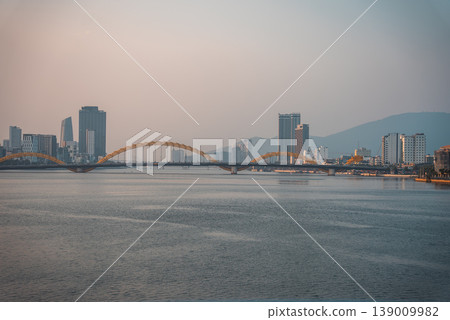 Dragon bridge over calm Han river at sunset with soft golden light and peaceful city skyline of Da Nang, tranquil urban landscape with serene water and warm evening atmosphere, Danang Vietnam Dragon bridge over calm Han river at sunset with soft golden light and peaceful city skyline of Da Nang, tranquil urban landscape with serene water and warm evening atmosphere, Danang Vietnam 139009982