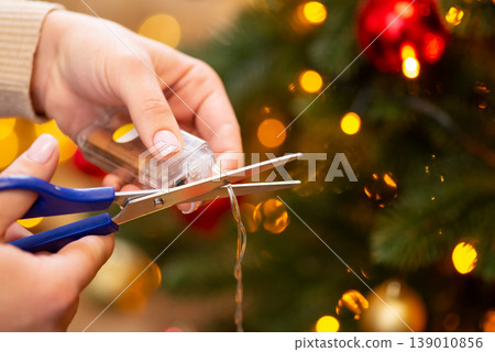 Woman holding socket with two AA batteries and scissors, trying to cut the wire of garland, close up. The end of winter holidays, finishing celebrations, spoiled festive mood concept Woman holding socket with two AA batteries and scissors, trying to cut the wire of garland, close up. The end of winter holidays, finishing celebrations, spoiled festive mood concept 139010856