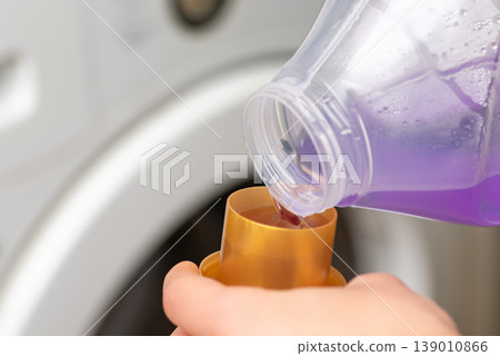 Liquid violet gel pouring into bottle cap in woman hands, close up. Concept of washing clothes, domestic routine and household chores Liquid violet gel pouring into bottle cap in woman hands, close up. Concept of washing clothes, domestic routine and household chores 139010866