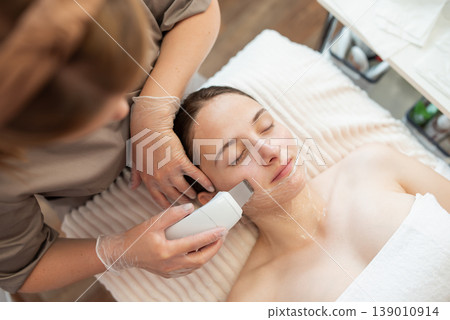 High angle view of young woman client lying with closed eyes and getting ultrasound skin cleaning. Cosmetology procedure in modern health and beauty salon 139010914