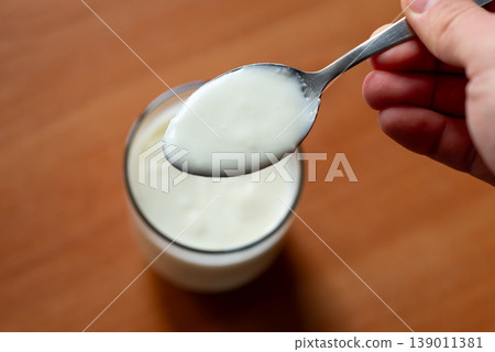 Liquid yogurt in spoon and glass in hand of a person, close up. Homemade dairy product standing on the wooden table Liquid yogurt in spoon and glass in hand of a person, close up. Homemade dairy product standing on the wooden table 139011381