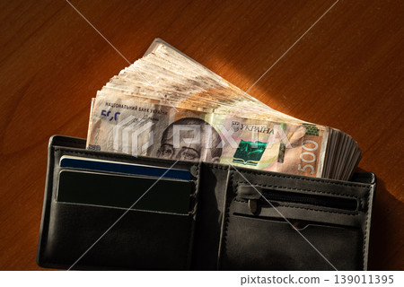 Many banknotes of 500 hryvnias in black wallet on the wooden table under the sunlight, close up. Ukrainian money, concept of finances, richness and good earnings Many banknotes of 500 hryvnias in black wallet on the wooden table under the sunlight, close up. Ukrainian money, concept of finances, richness and good earnings 139011395