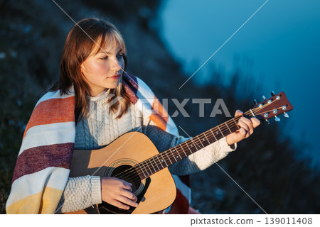 Portrait of a young girl playing the guitar near the river. Girl sitting on river bank and practising playing the guitar, mastering skills, learning new composition 139011408