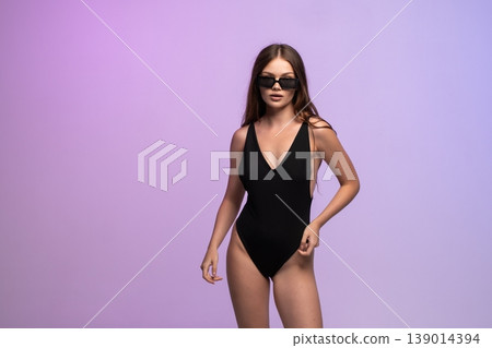 Confident young woman posing in black swimsuit and stylish sunglasses on purple gradient background 139014394