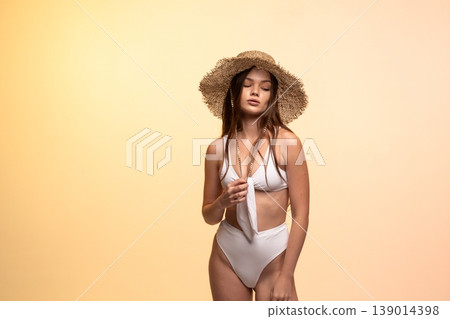 Young woman wearing a white bikini and straw sun hat, posing with eyes closed embracing summer fashion and relaxation 139014398