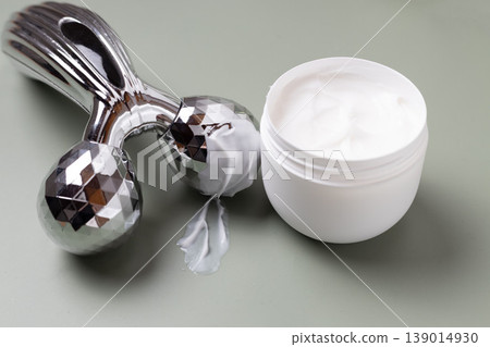 Metal facial roller with moisturizing skincare cream and cosmetic jar on neutral background, beauty treatment concept 139014930