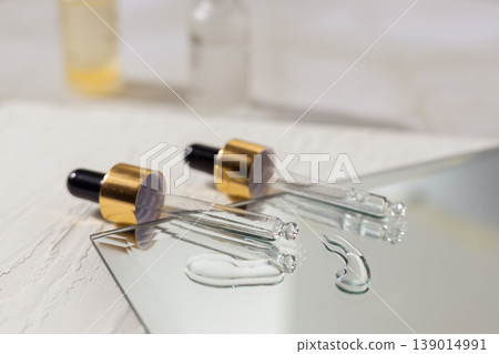 Glass cosmetic droppers with transparent serum on mirror surface, skincare essence drops and reflection, beauty treatment concept, minimalist cosmetic product background. Glass cosmetic droppers with transparent serum on mirror surface, skincare essence drops and reflection, beauty treatment concept, minimalist cosmetic product background. 139014991