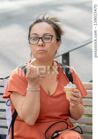 Woman age 45 eats ice cream on a bench during a city walk in summer. Woman age 45 eats ice cream on a bench during a city walk in summer. 139015470