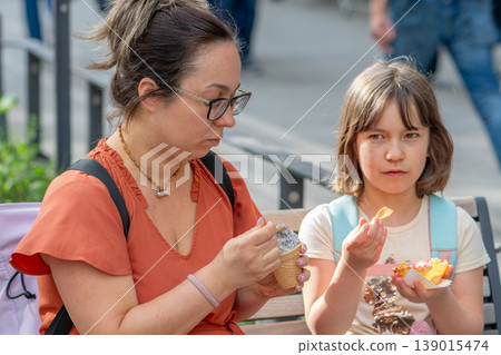 Mother and daughter eat ice cream on a bench during a walk through the city. Mother and daughter eat ice cream on a bench during a walk through the city. 139015474