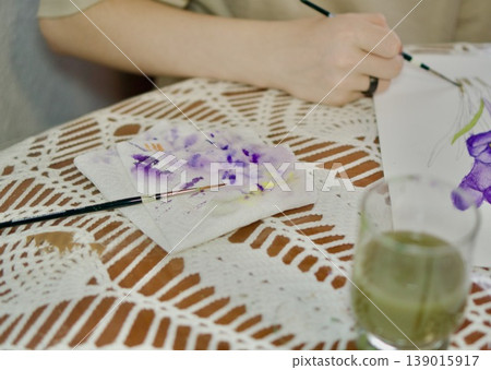 Artist's hands painting purple crocuses with watercolors. Creative workspace with palette and brushes, art education concept. Artist's hands painting purple crocuses with watercolors. Creative workspace with palette and brushes, art education concept. 139015917