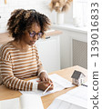 female architect designer is working, taking notes in notebook is mock up of house on table. 139016833
