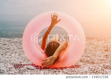 Beach summer vacation woman relaxes on a pebble shore with a large pink inflatable ring during a warm golden hour sunset Beach summer vacation woman relaxes on a pebble shore with a large pink inflatable ring during a warm golden hour sunset 139017198
