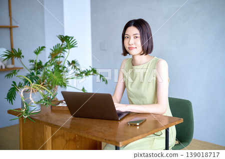 A portrait of an Asian woman smiling and looking at the camera while using her laptop at home. A portrait of an Asian woman smiling and looking at the camera while using her laptop at home. 139018717